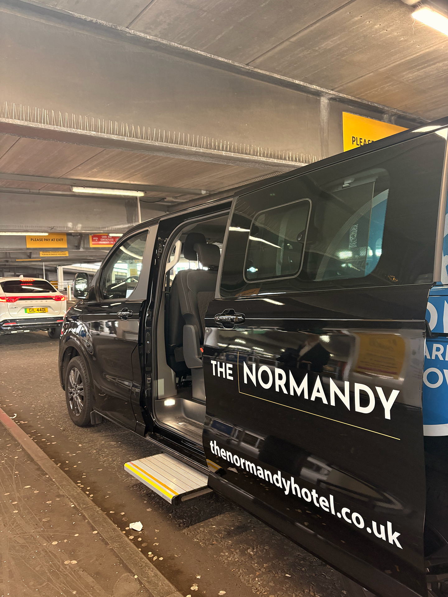 The Normandy Hotel and Zoom Airport Parking executive shuttle minibus at the Glasgow Airport passenger pickup zone with the side door open.