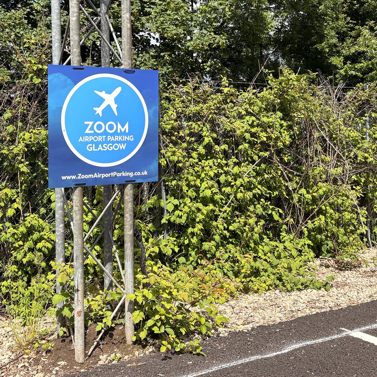 Square blue Zoom Airport Parking Glasgow branded sign mounted on a steel pillar next to a secure wire fence and green hedges.