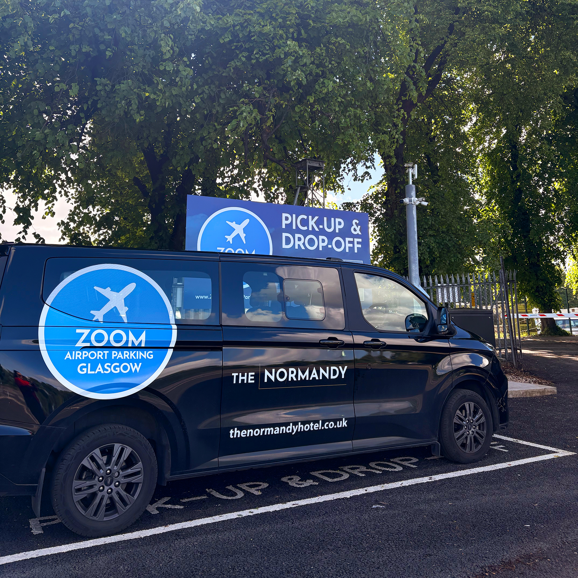 Zoom Airport Parking Glasgow branded shuttle van parked in the designated pick-up and drop-off zone with a security CCTV pole and trees in the background.